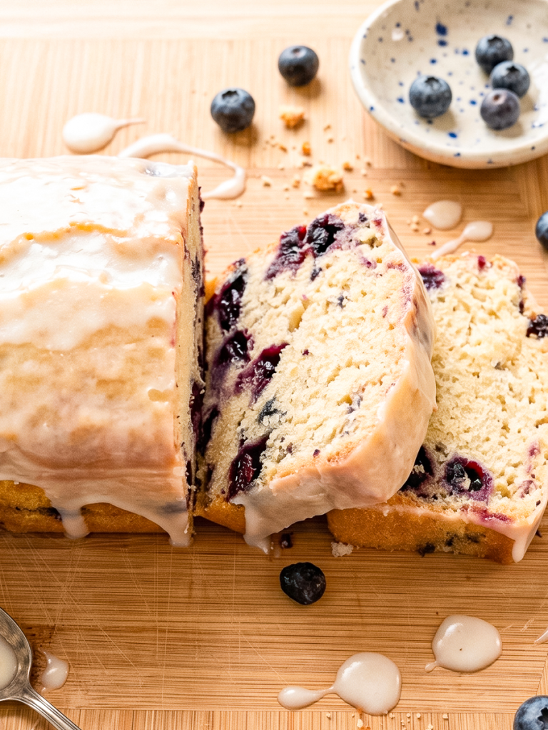 A glazed blueberry lemon loaf, partially sliced to show the fruit inside, rests on a wooden board with scattered blueberries and crumbs.