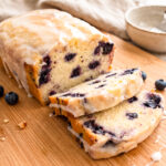 A glazed blueberry lemon loaf, partially sliced, on a wooden board with scattered blueberries and glaze drips.