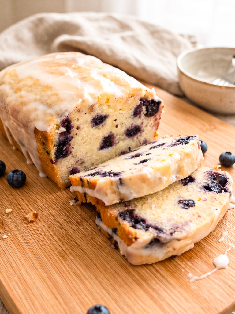 A glazed blueberry lemon loaf, partially sliced, on a wooden board with scattered blueberries and glaze drips.