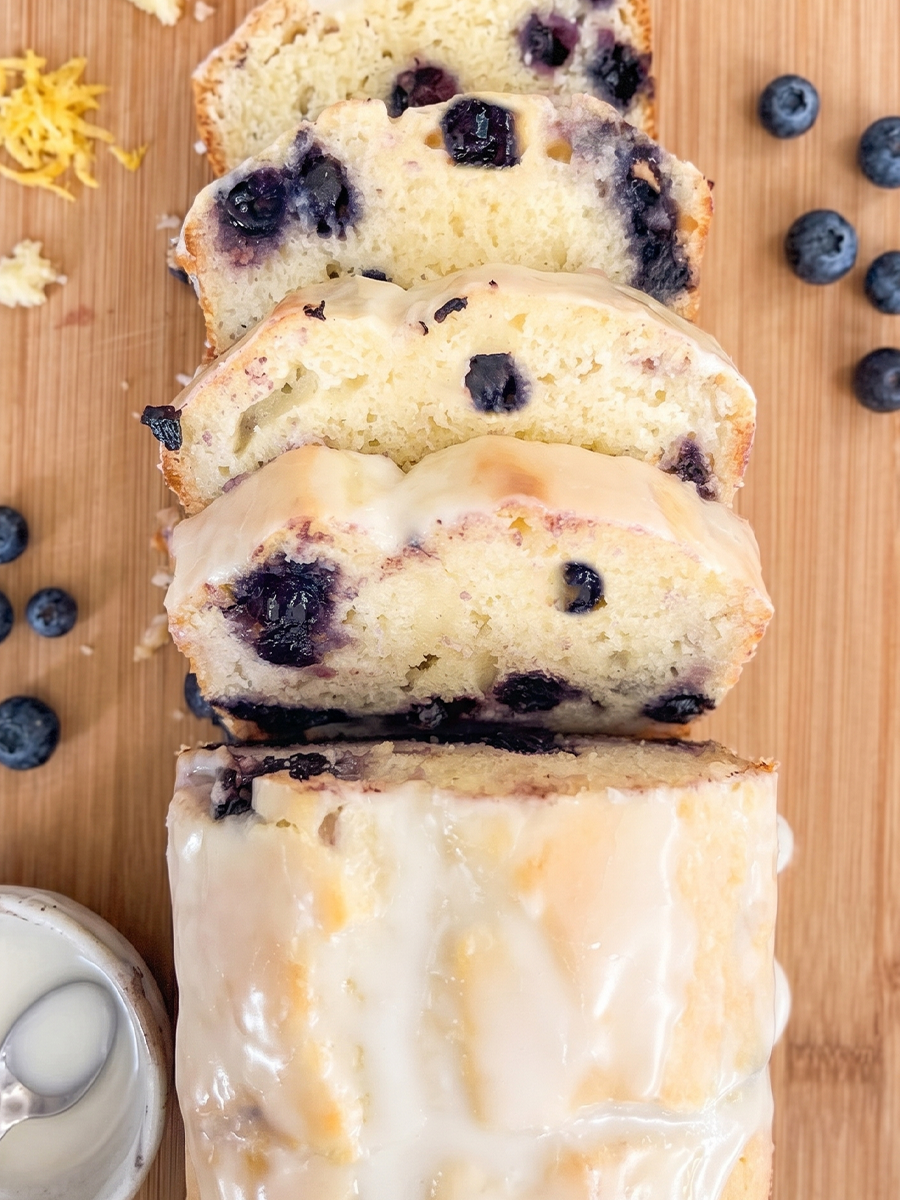 A glazed blueberry lemon loaf is sliced on a wooden board, with fresh blueberries, lemon zest, and a bowl of glaze nearby.