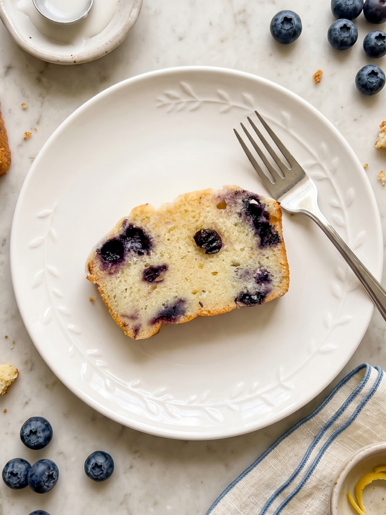 A slice of blueberry lemon loaf cake with a fork on a white plate, surrounded by fresh blueberries, a napkin, and lemon zest on a marble surface.