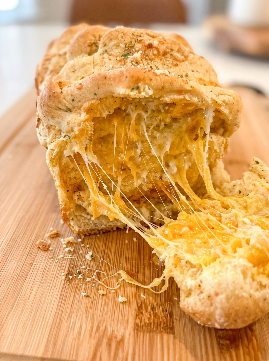 A warm loaf of pull-apart bread on a wooden board shows gooey, melted orange cheese stretching as a piece is pulled away.