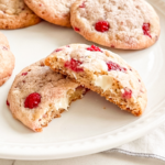 Several raspberry white chocolate cookies are on a white plate, with one broken in half revealing its soft interior and melted white chocolate.