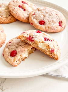 Several raspberry white chocolate cookies are on a white plate, with one broken in half revealing its soft interior and melted white chocolate.