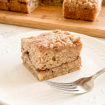 A slice of banana coffee cake with a streusel topping and cinnamon swirl is on a white plate with a fork; more cake is on a wooden board.