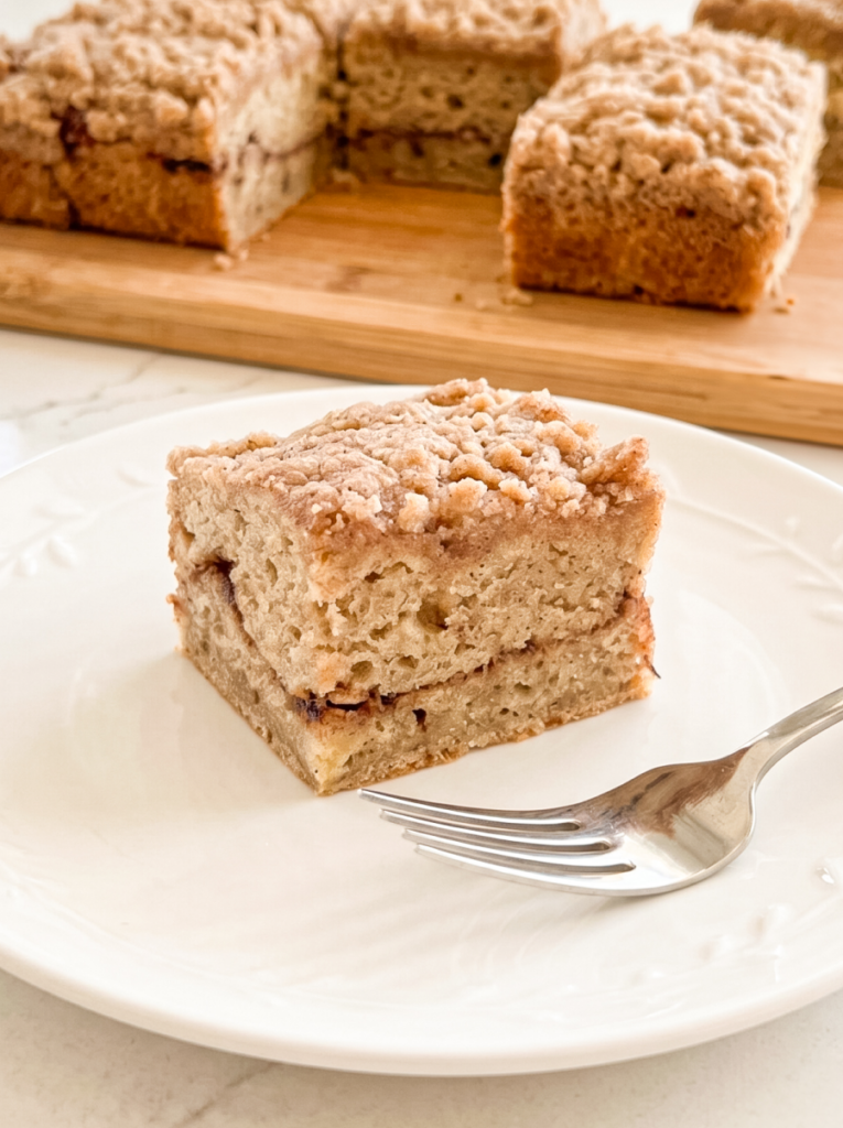 A slice of banana coffee cake with a streusel topping and cinnamon swirl is on a white plate with a fork; more cake is on a wooden board.