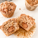 Three biscoff streusel banana muffins with crumbly topping are displayed on a marble surface, one cut in half revealing a creamy Biscoff center.