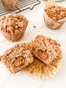 Three biscoff streusel banana muffins with crumbly topping are displayed on a marble surface, one cut in half revealing a creamy Biscoff center.