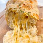 Golden garlic cheddar parm pull apart bread on a cutting board with melted cheese stretching out as a piece is pulled away.