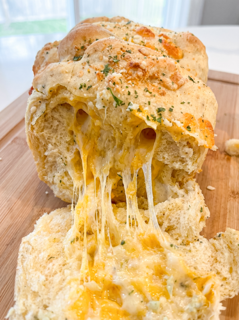 Golden garlic cheddar parm pull apart bread on a cutting board with melted cheese stretching out as a piece is pulled away.