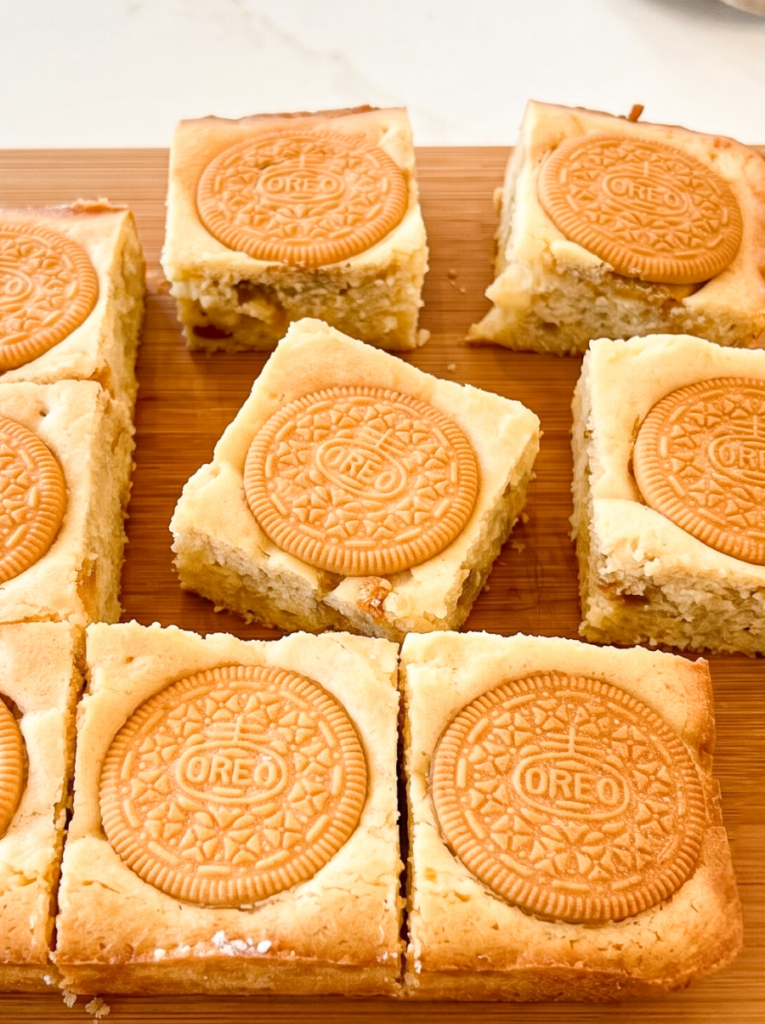 Square blondies with golden Oreo cookies on top are arranged on a wooden board.