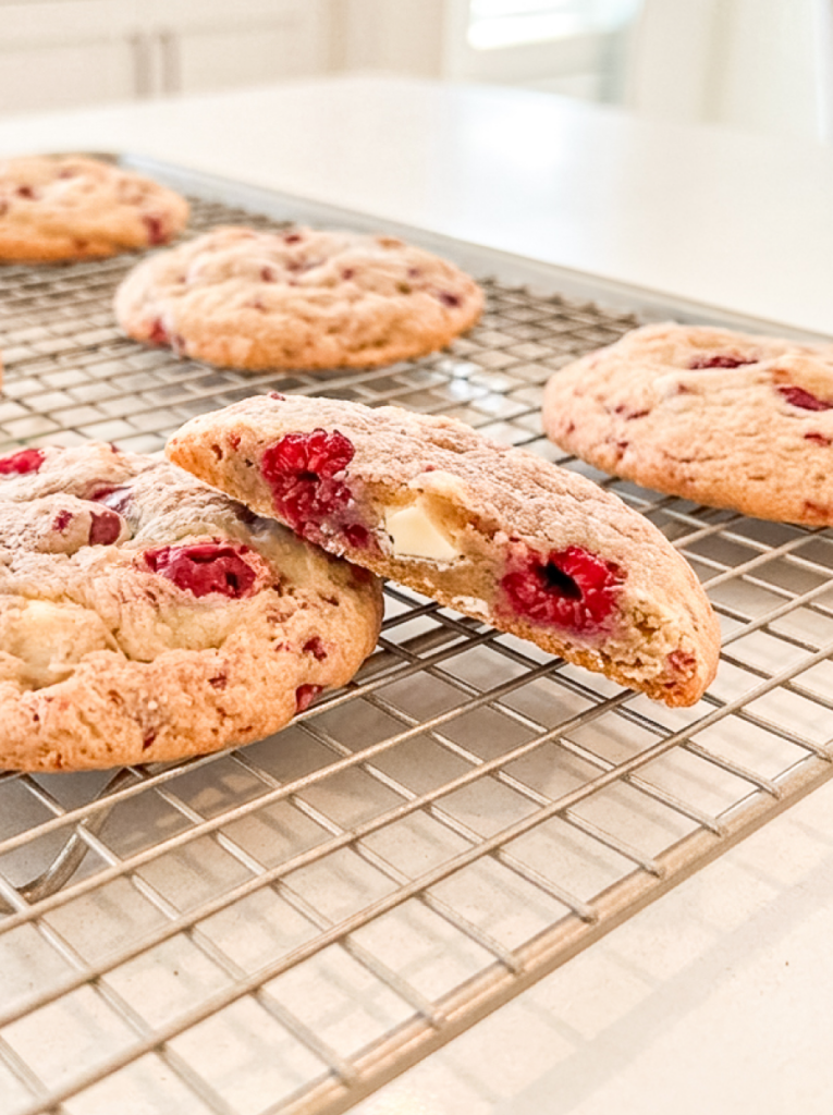 Freshly baked raspberry white chocolate cookies cooling on a wire rack, with one split open to show the fruit and chocolate.