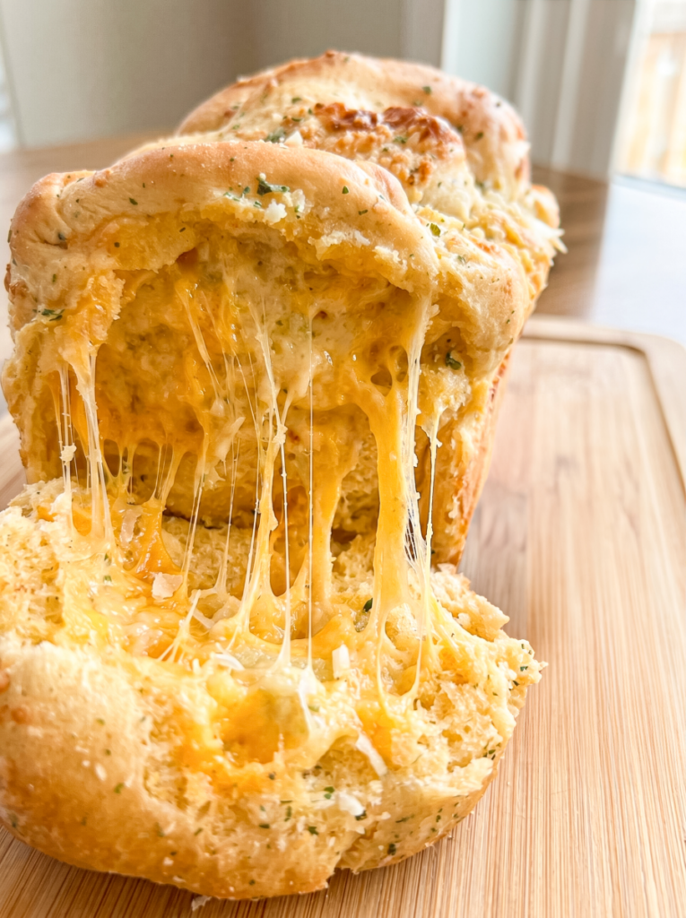 A golden-brown loaf of garlic cheddar parm pull-apart bread with gooey melted cheese stretching between the torn sections on a wooden board.