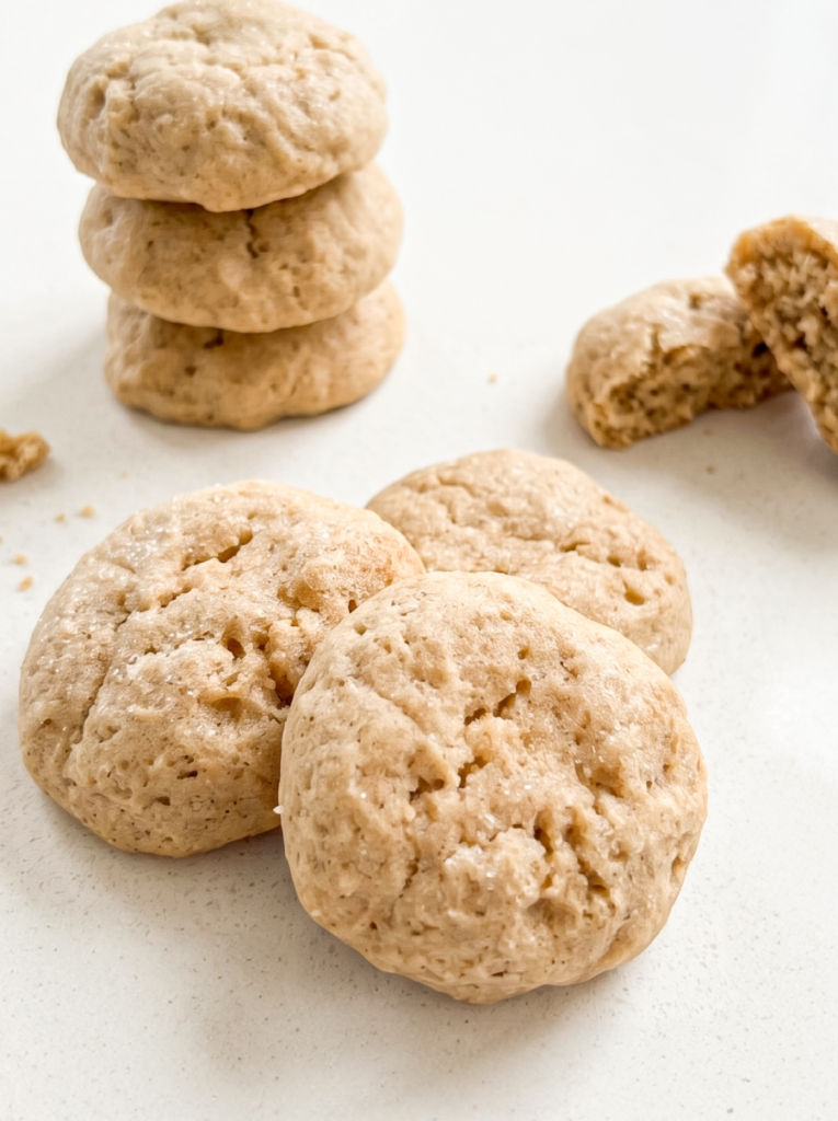 A stack of three light brown cookies and several individual cookies are on a white surface, with one broken in half to show its soft interior.