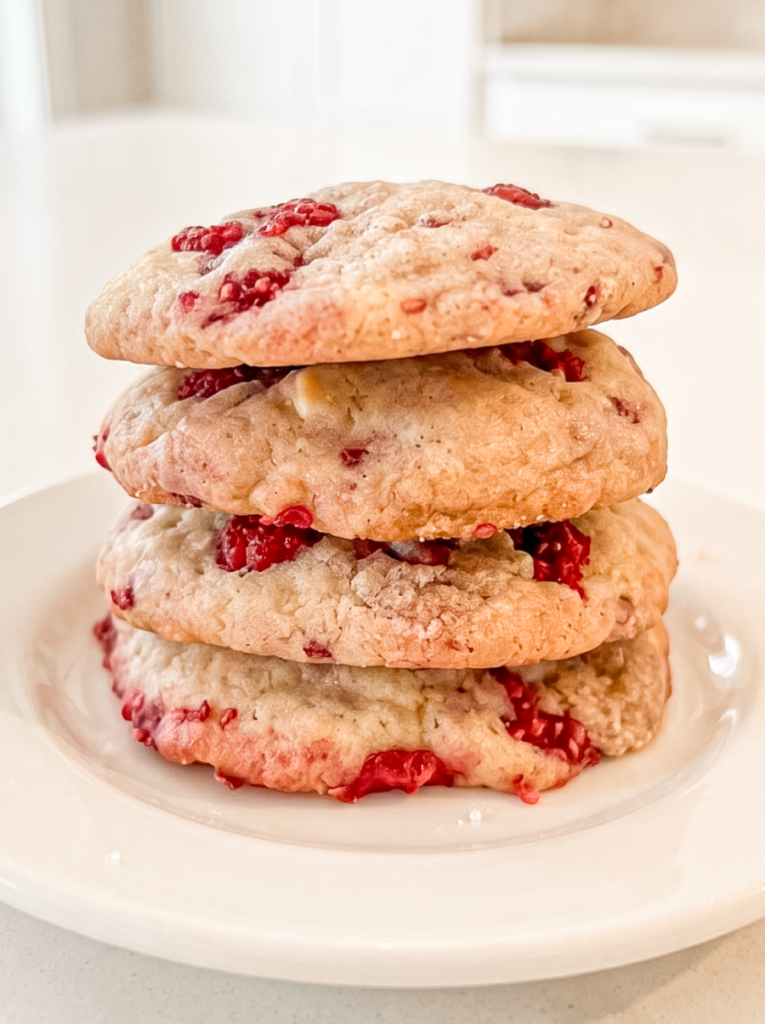 A stack of four golden-brown cookies with visible red raspberries and white chocolate chips rests on a white plate.