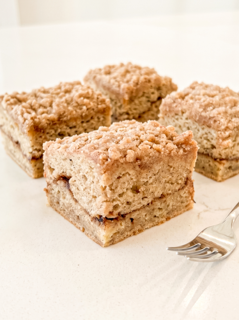Four square slices of banana coffee cake with a crumb topping and cinnamon swirl, next to a silver fork on a white surface.