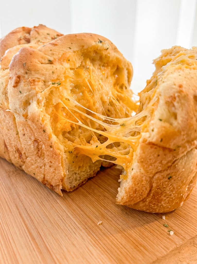 A golden-brown loaf of garlic cheddar parm pull-apart bread sits on a wooden board, with gooey melted cheese stretching from the middle.