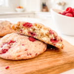 A raspberry white chocolate cookie is split on a wooden board, revealing melted white chocolate and raspberries inside, with whole cookies and fresh raspberries nearby.