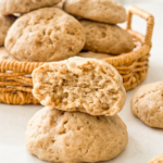 Soft banana cookies are piled in a woven basket, with one broken cookie revealing its fluffy interior in the foreground.
