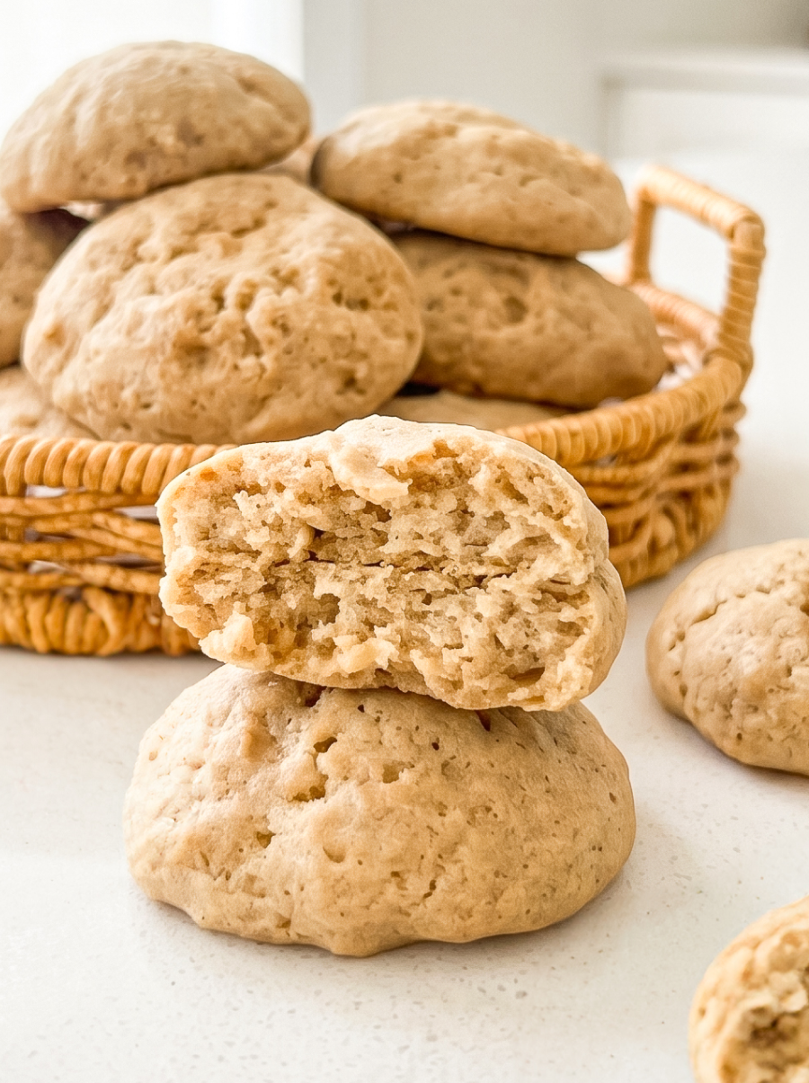 Soft banana cookies are piled in a woven basket, with one broken cookie revealing its fluffy interior in the foreground.