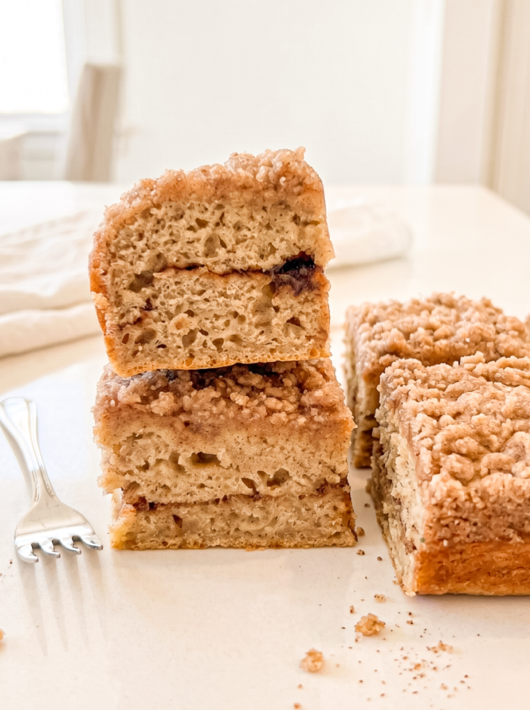 Two squares of banana coffee cake are stacked to show their airy texture and cinnamon swirl, with a streusel topping, a fork, and more cake.