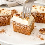A fork punctures a square slice of carrot cake topped with thick cream cheese frosting and walnuts on a white plate.