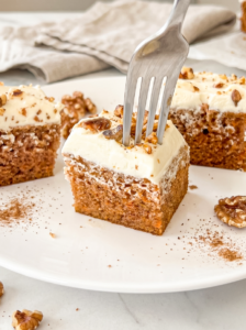 A fork punctures a square slice of carrot cake topped with thick cream cheese frosting and walnuts on a white plate.