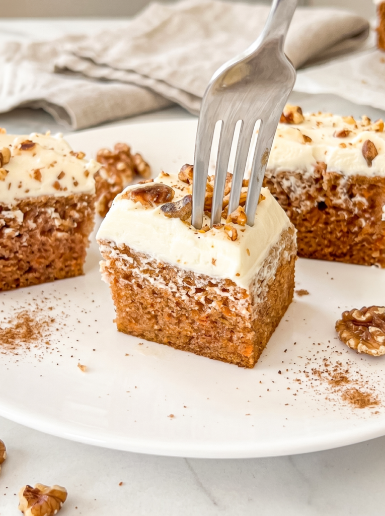 A fork punctures a square slice of carrot cake topped with thick cream cheese frosting and walnuts on a white plate.