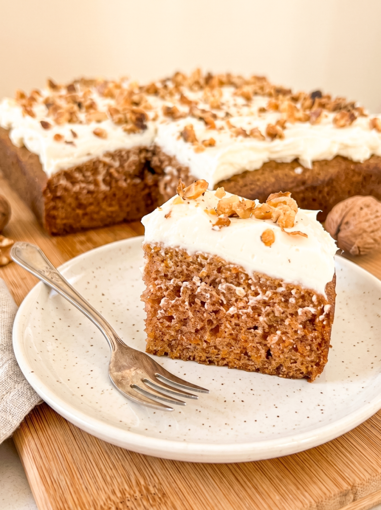 A slice of carrot sheet cake with thick cream cheese frosting and walnuts sits on a plate with a fork, with the full cake in the background.
