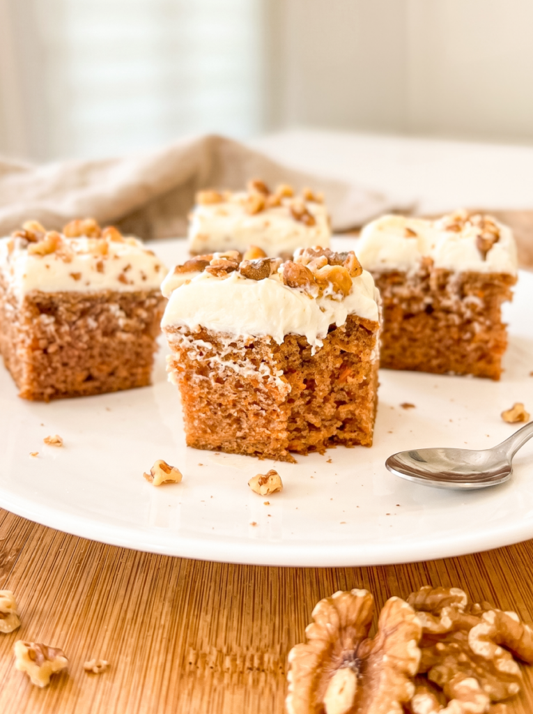 Four square carrot cake pieces with thick cream cheese frosting and walnuts on a white plate, with a spoon and scattered walnuts.