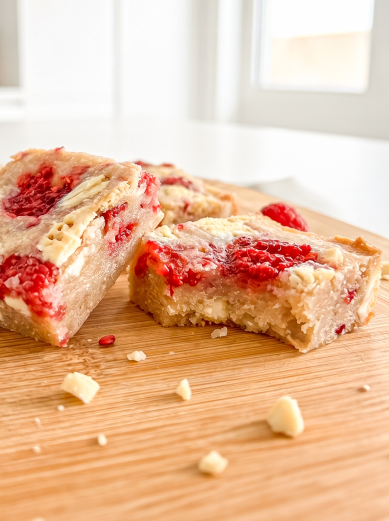 Two raspberry white chocolate blondies, one with a bite taken, sit on a wooden cutting board with scattered white chocolate chips.