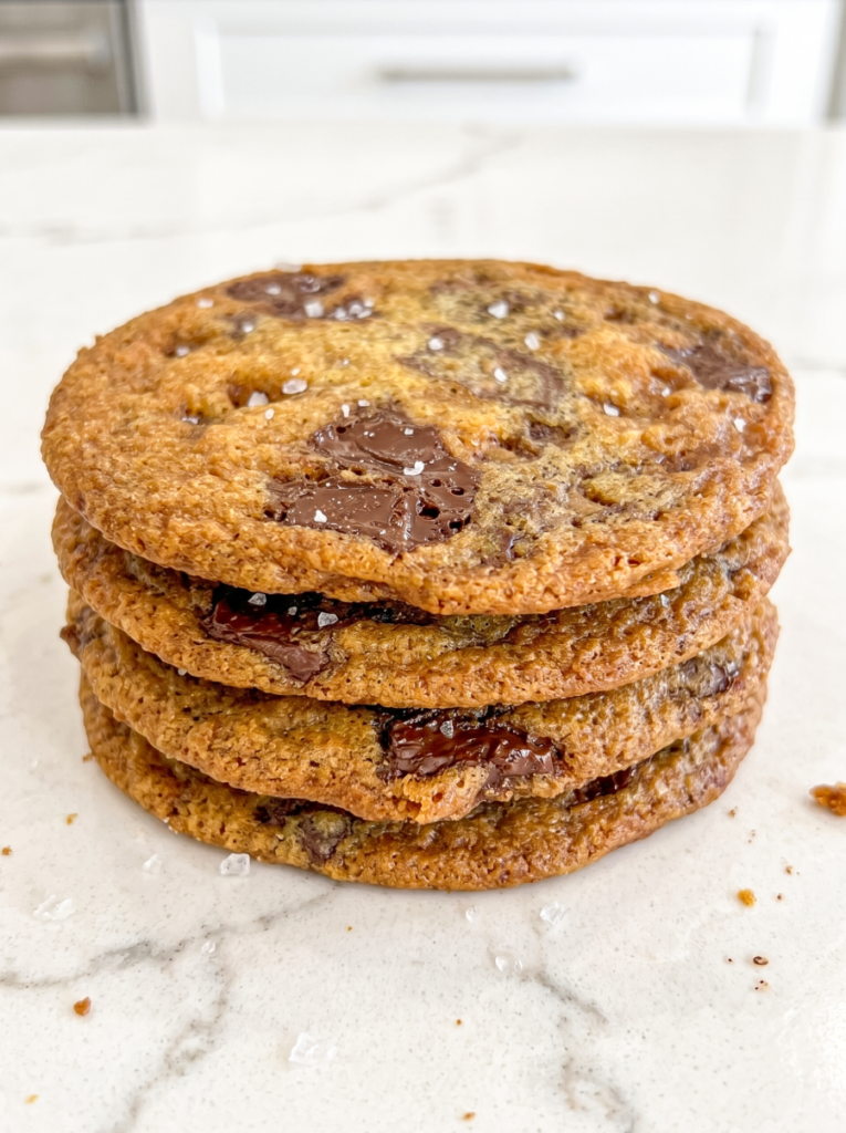 A stack of four golden-brown cookies with melted chocolate chunks and sea salt flakes on a white marble surface.