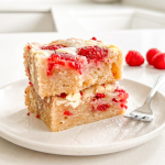 Two raspberry white chocolate blondies are stacked on a white plate with a fork and fresh raspberries in the bright kitchen background.