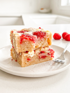 Two raspberry white chocolate blondies are stacked on a white plate with a fork and fresh raspberries in the bright kitchen background.