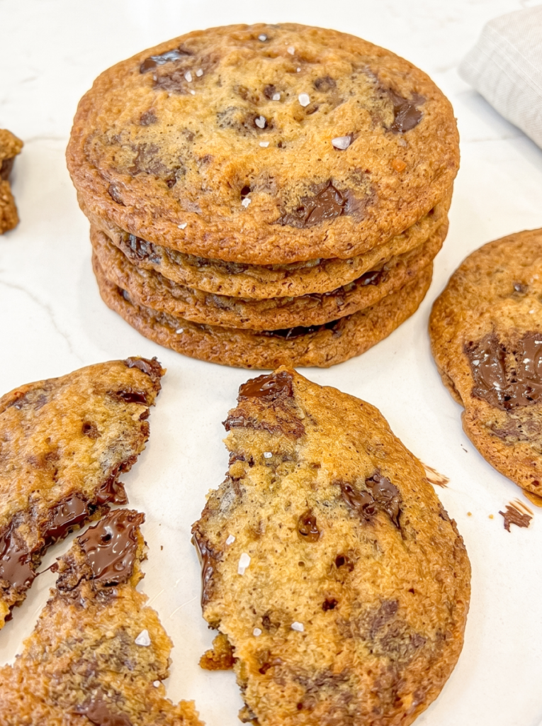 A stack of golden-brown chocolate toffee cookies with sea salt, next to a broken cookie revealing melted chocolate on a marble surface.