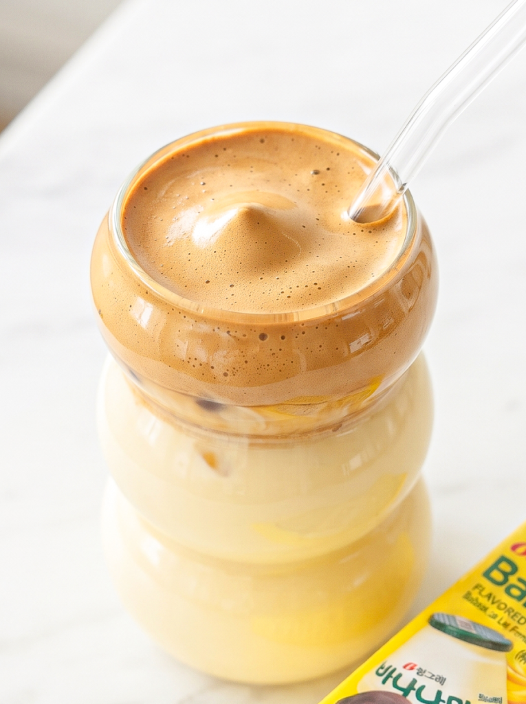 A clear glass with yellow banana milk and brown dalgona coffee foam, with a straw and a yellow banana milk carton visible.