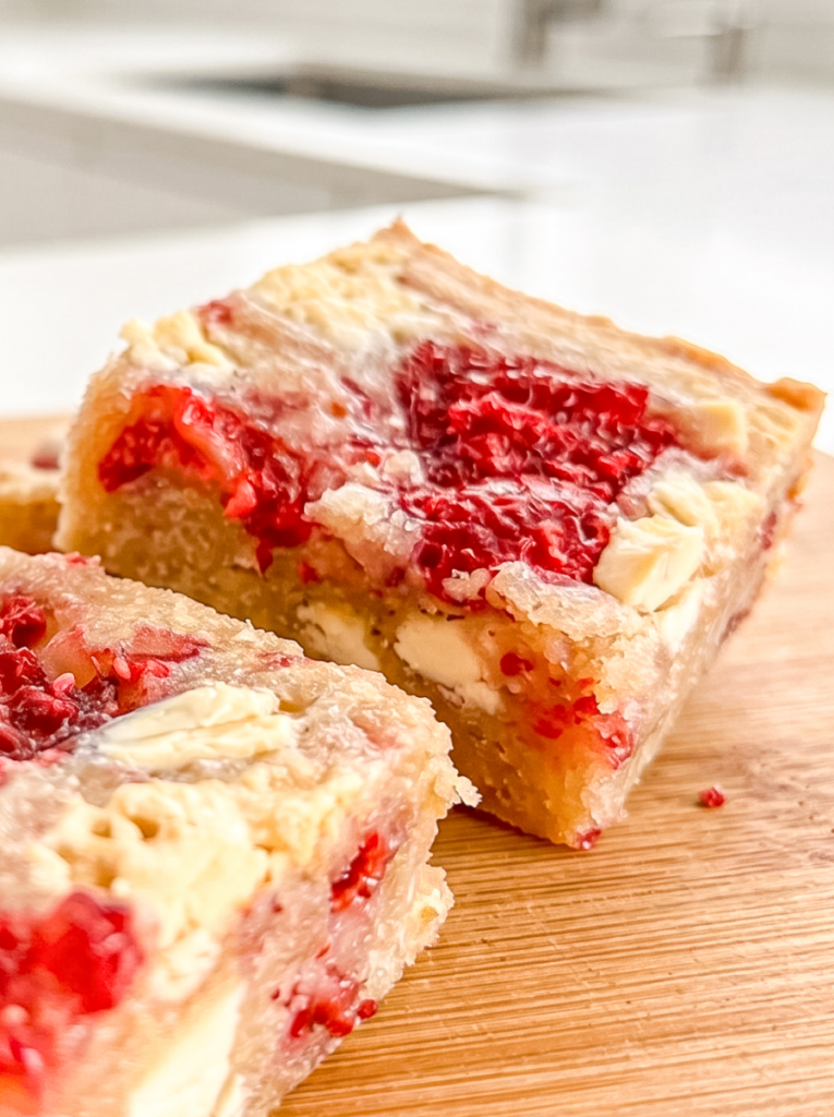 Two raspberry white chocolate blondies, one large and one smaller, sit on a wooden board, showcasing their vibrant raspberries and white chocolate.