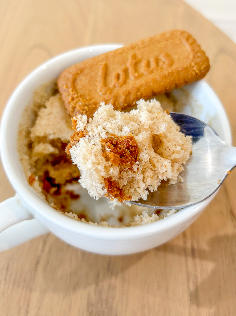 A spoon scoops a fluffy piece of biscoff mug cake from a white mug, topped with a Lotus cookie, on a wooden table.