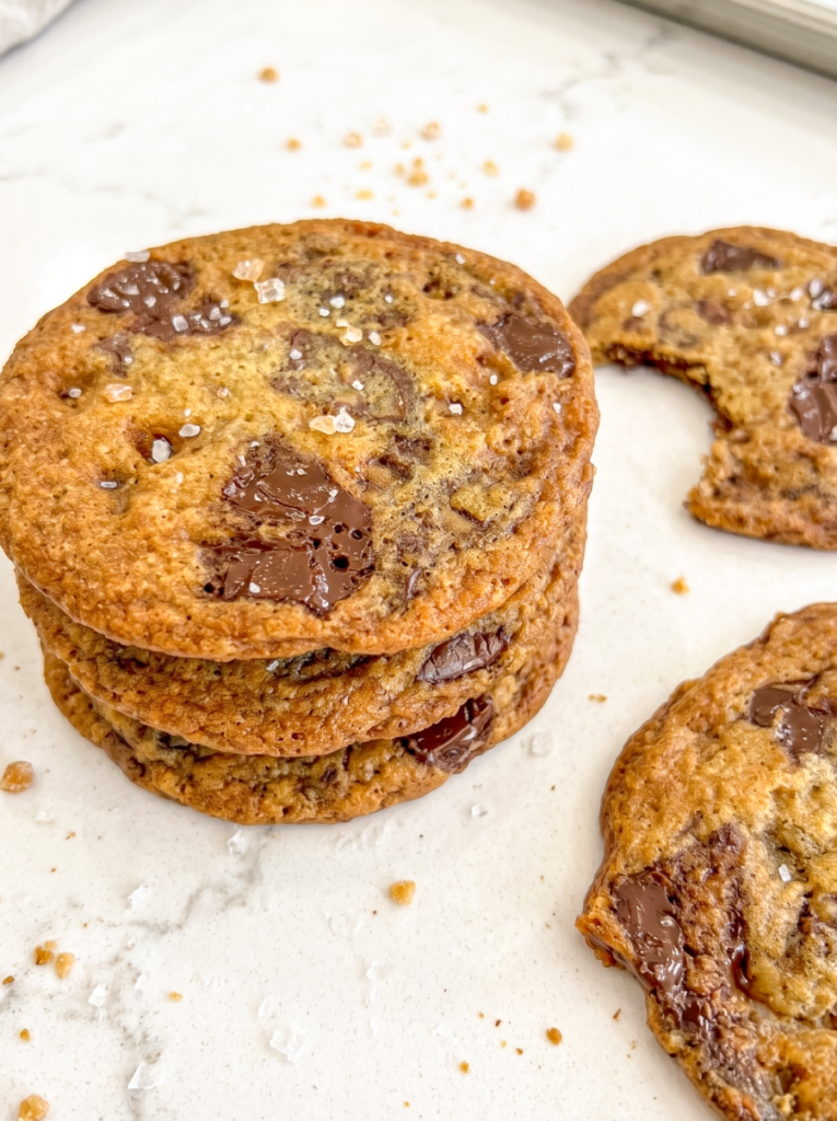 A stack of golden-brown chocolate toffee cookies with sea salt, next to a broken cookie revealing melted chocolate on a marble surface.