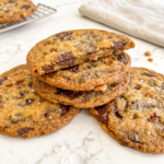 A stack of three golden-brown chocolate toffee cookies with flaky salt, the top one broken to show its gooey center, on a marble counter.