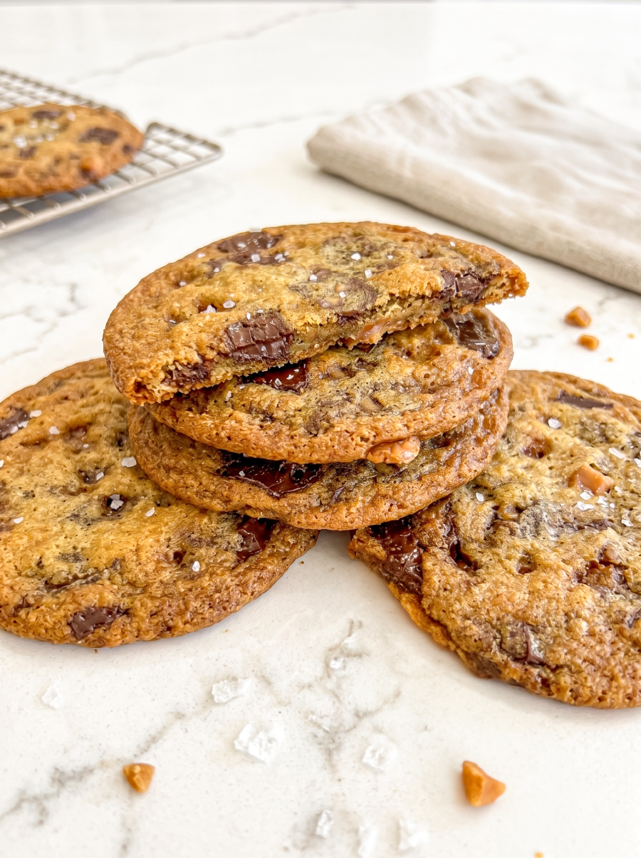 A stack of three golden-brown chocolate toffee cookies with flaky salt, the top one broken to show its gooey center, on a marble counter.