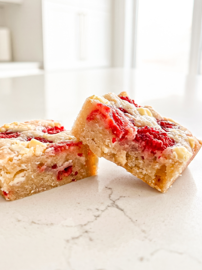 Two raspberry white chocolate blondies with red fruit swirls and white chips sit on a light countertop in a bright kitchen.