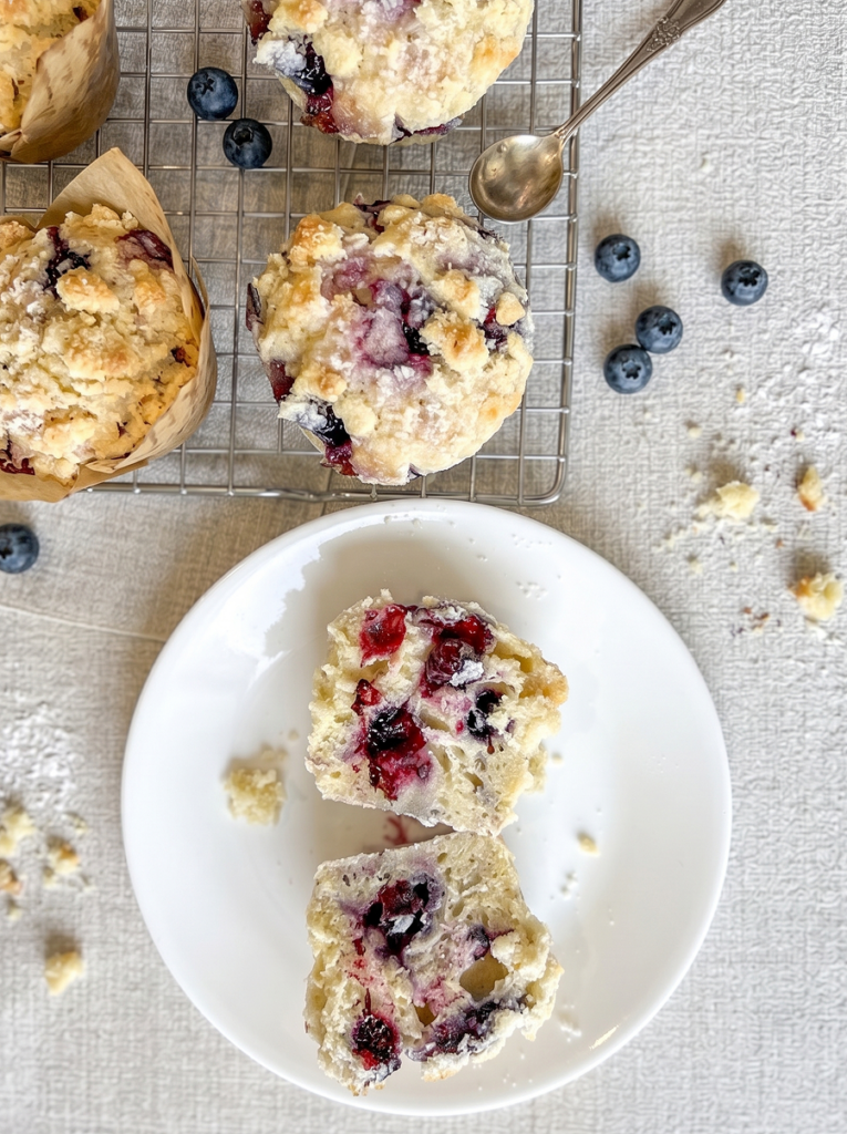 Bakery style blueberry muffins with streusel topping, some on a cooling rack and one cut open on a plate to show the juicy blueberries.