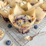Freshly baked blueberry muffins with streusel topping in tulip liners sit on a cooling rack with loose blueberries and a spoon.