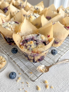 Freshly baked blueberry muffins with streusel topping in tulip liners sit on a cooling rack with loose blueberries and a spoon.