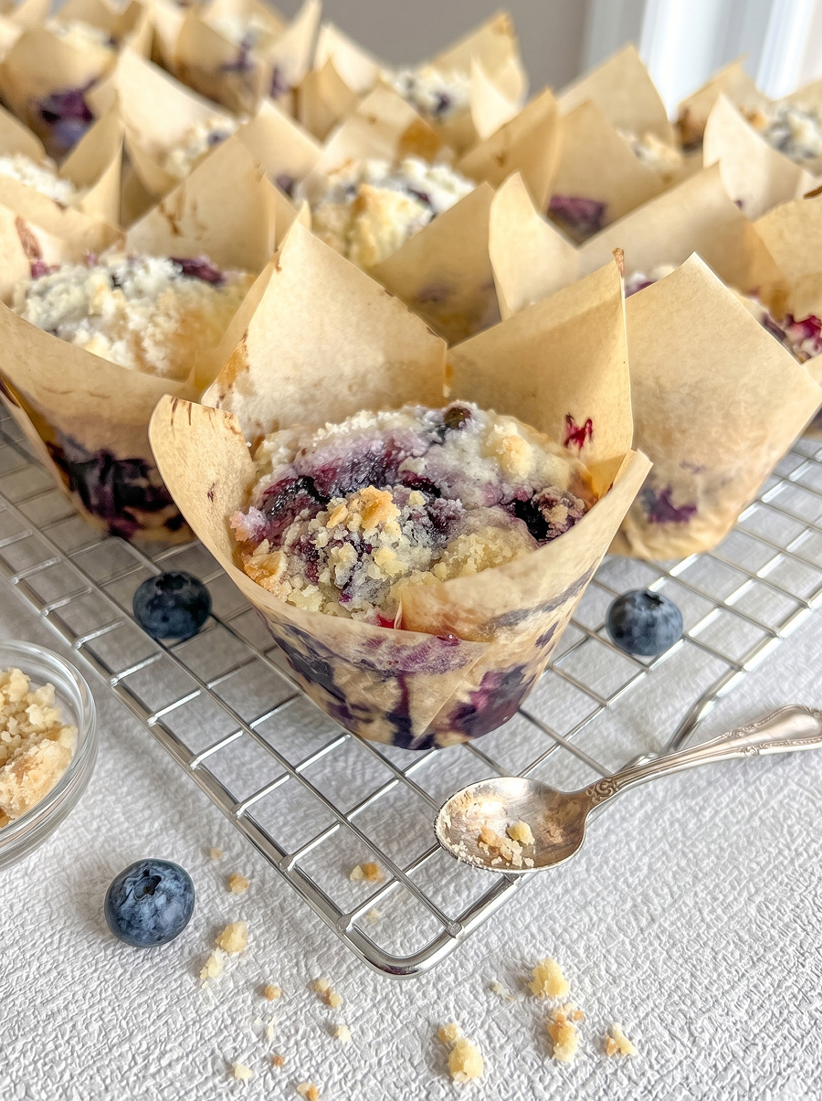 Freshly baked blueberry muffins with streusel topping in tulip liners sit on a cooling rack with loose blueberries and a spoon.