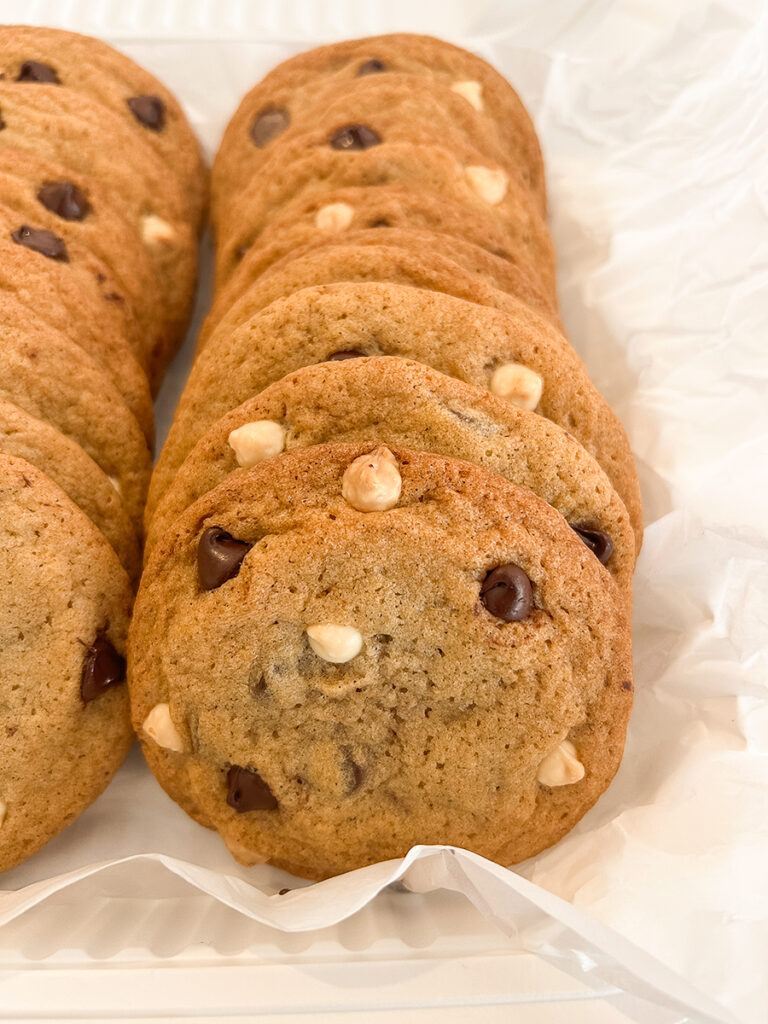 Rows of golden-brown cookies with dark and white chocolate chips are arranged on white parchment paper in a tray.