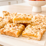 Several peach crumble bars with a golden peach filling, crumbly topping, and a light glaze, arranged on a white board with fresh peaches in the background.