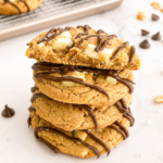 A stack of four peanut butter cookies with white chocolate chunks and dark chocolate drizzle on a white marble surface, with one cookie broken.