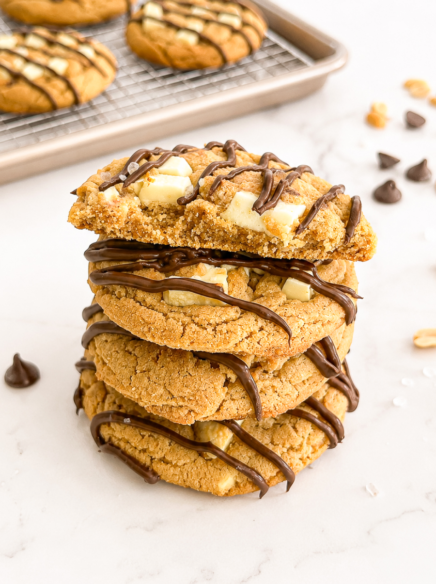 A stack of four peanut butter cookies with white chocolate chunks and dark chocolate drizzle on a white marble surface, with one cookie broken.